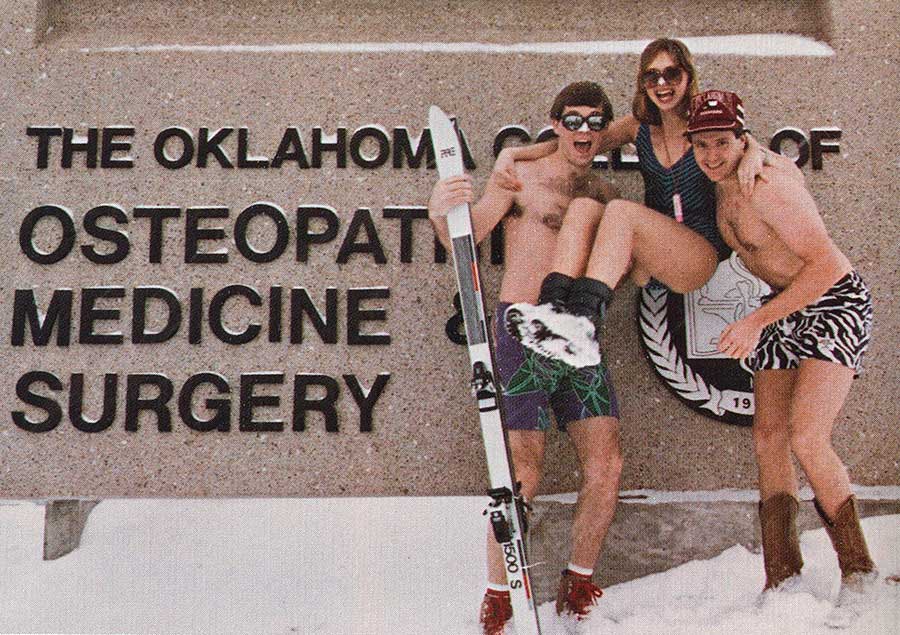 3 students posing in snow while wearing swim wear as a prank in front of the school's sign